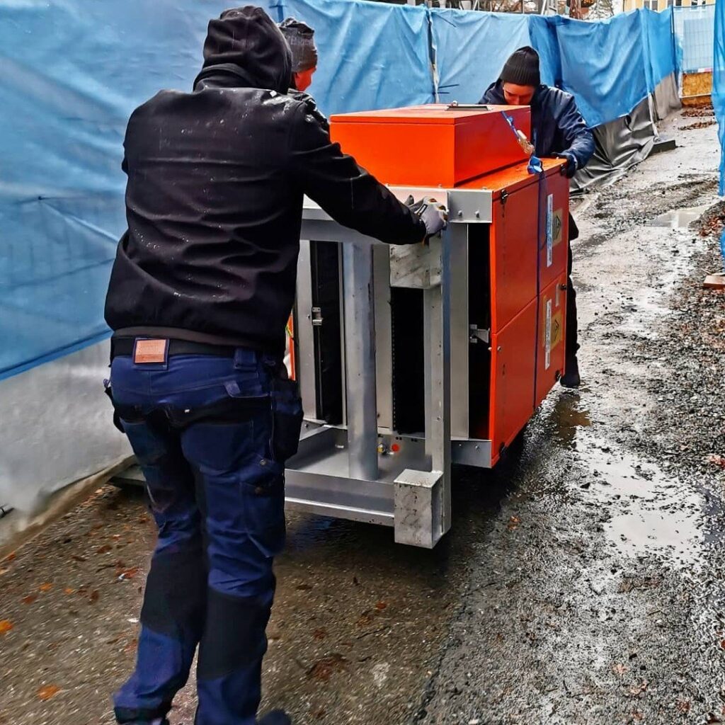 Två män skjuter ett orange aggregat på stålram längs en blöt grusväg på en byggplats