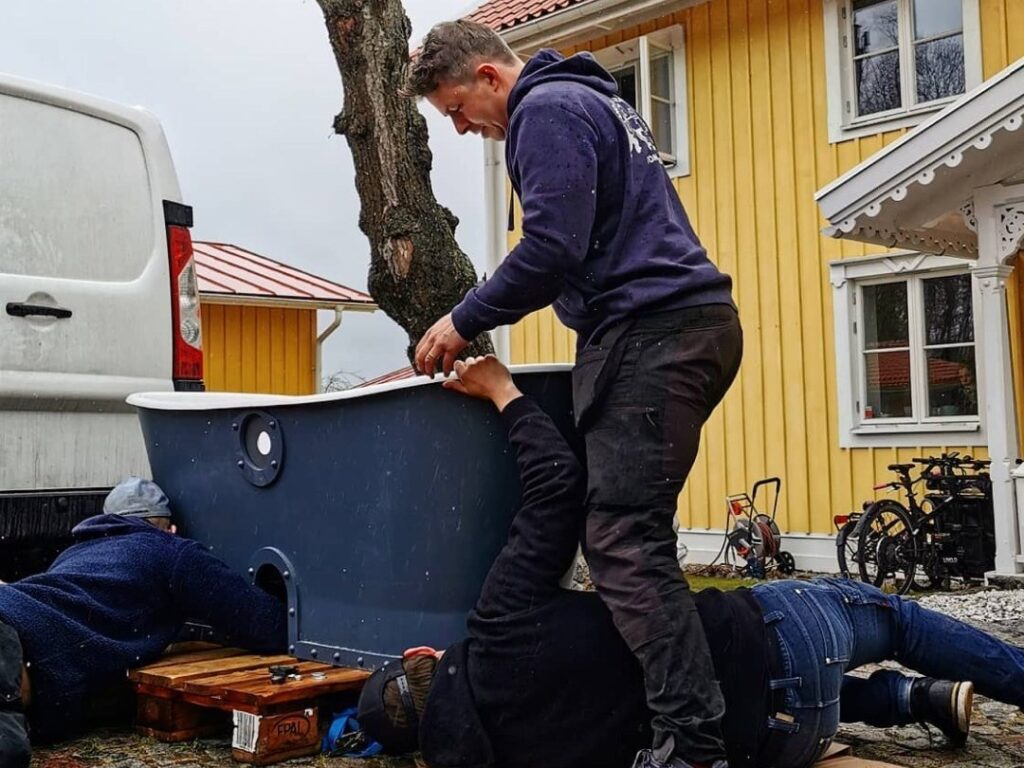 Tre personer lyfter ett mörkblått gjutjärnsbadkar från en lastpall framför ett gult trähus